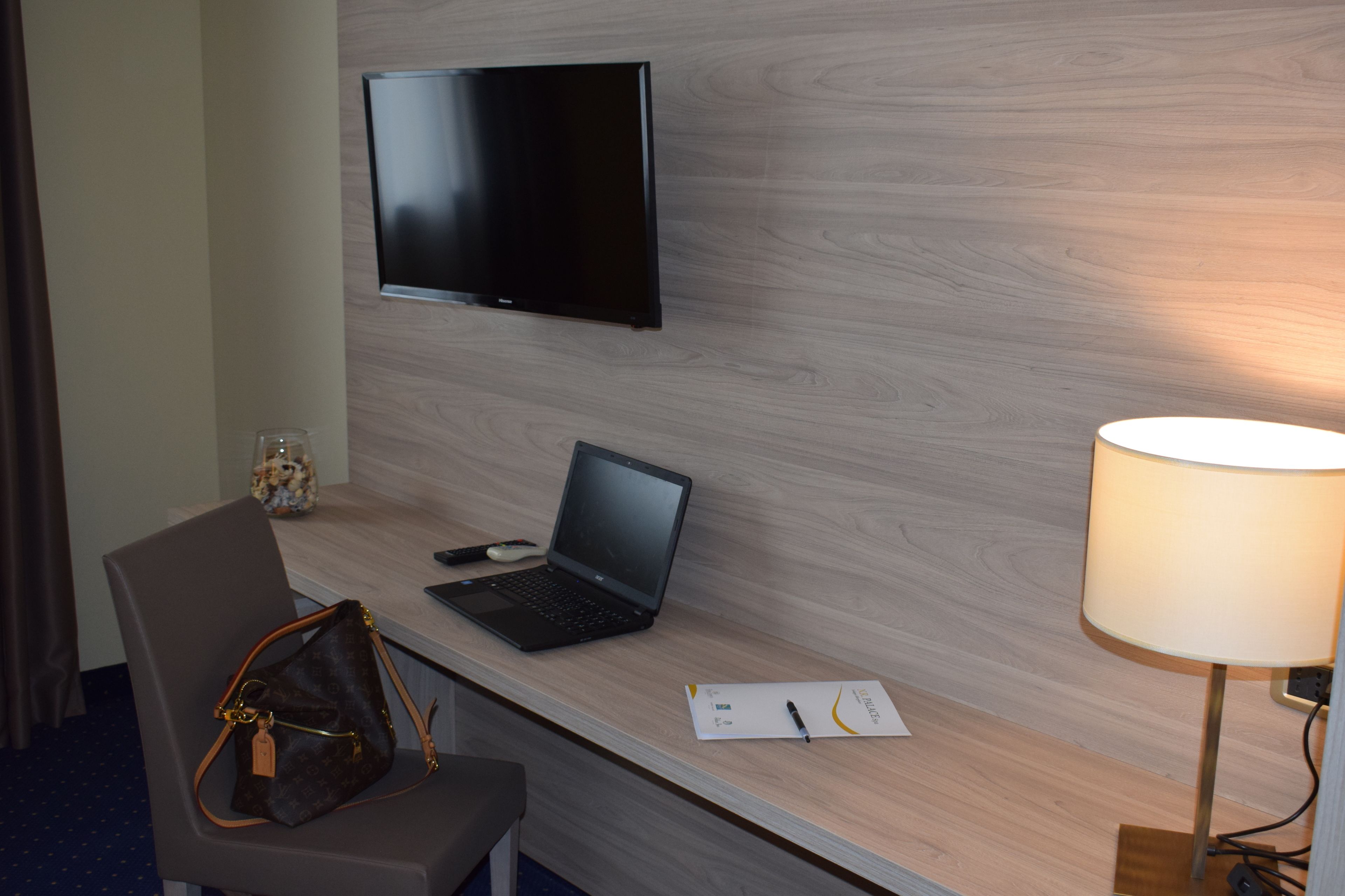 Habitación de hotel con un escritorio de madera, una computadora portátil, un televisor montado en la pared, una lámpara y una silla.