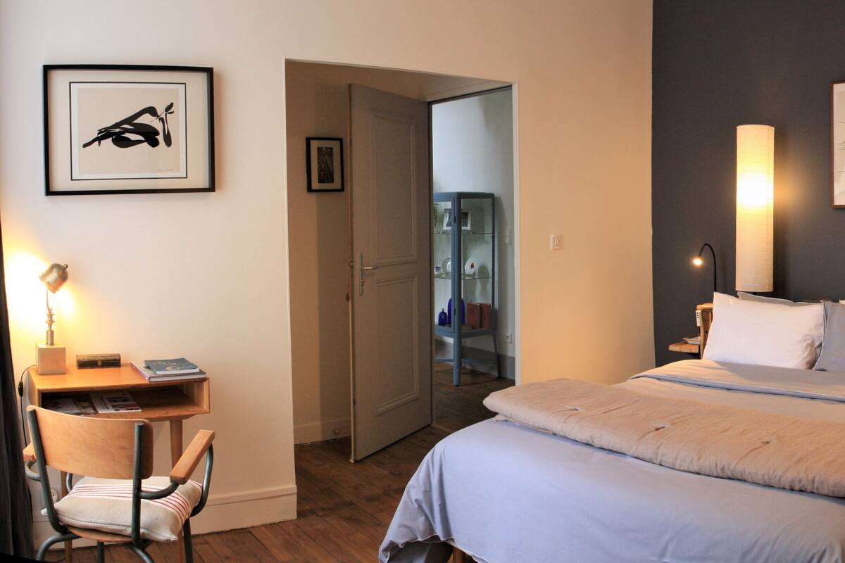 A bedroom with a bed, bedside tables, a desk, and a chair. There is a door leading to another room.