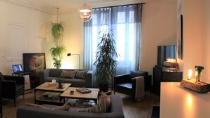 A modern living room with a sofa, coffee table, and two armchairs. There are potted plants, a lamp, and a small table with decorative items. - La Villa Dunois (Orléans)