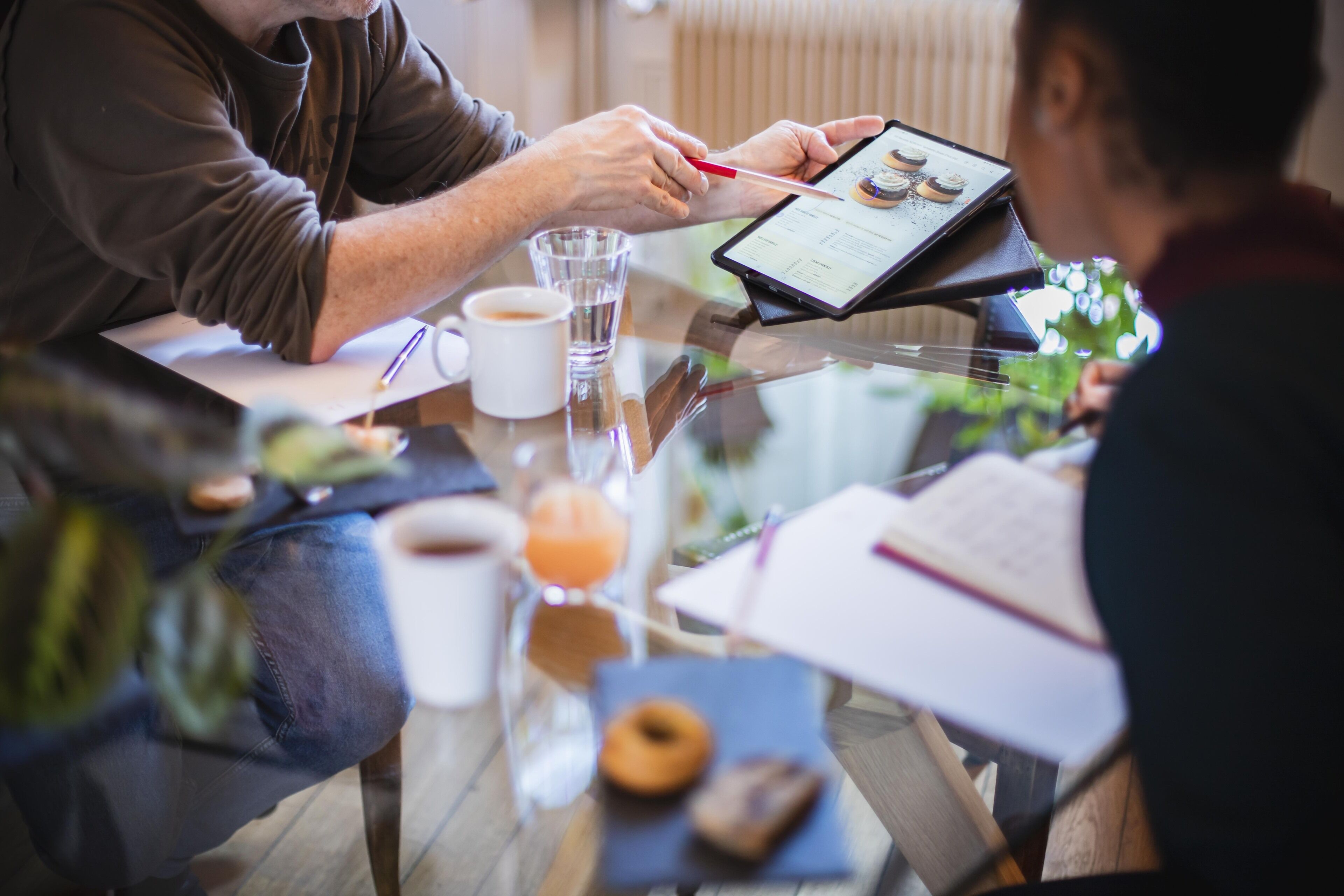 Une personne utilise une tablette à une table où se trouvent un verre d’eau, une tasse de café et un beignet.