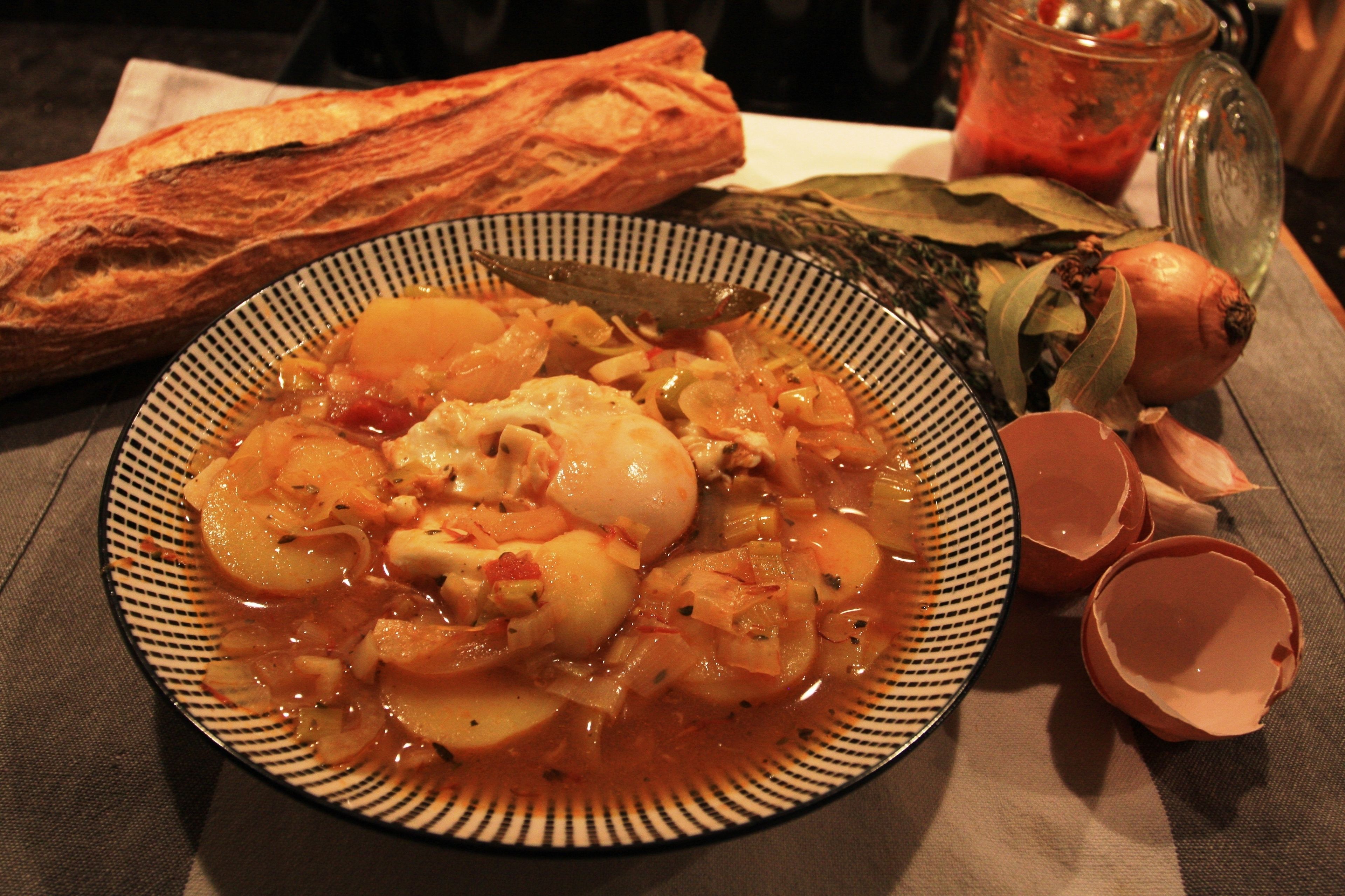 A plate of soup with vegetables and a baguette on a table.