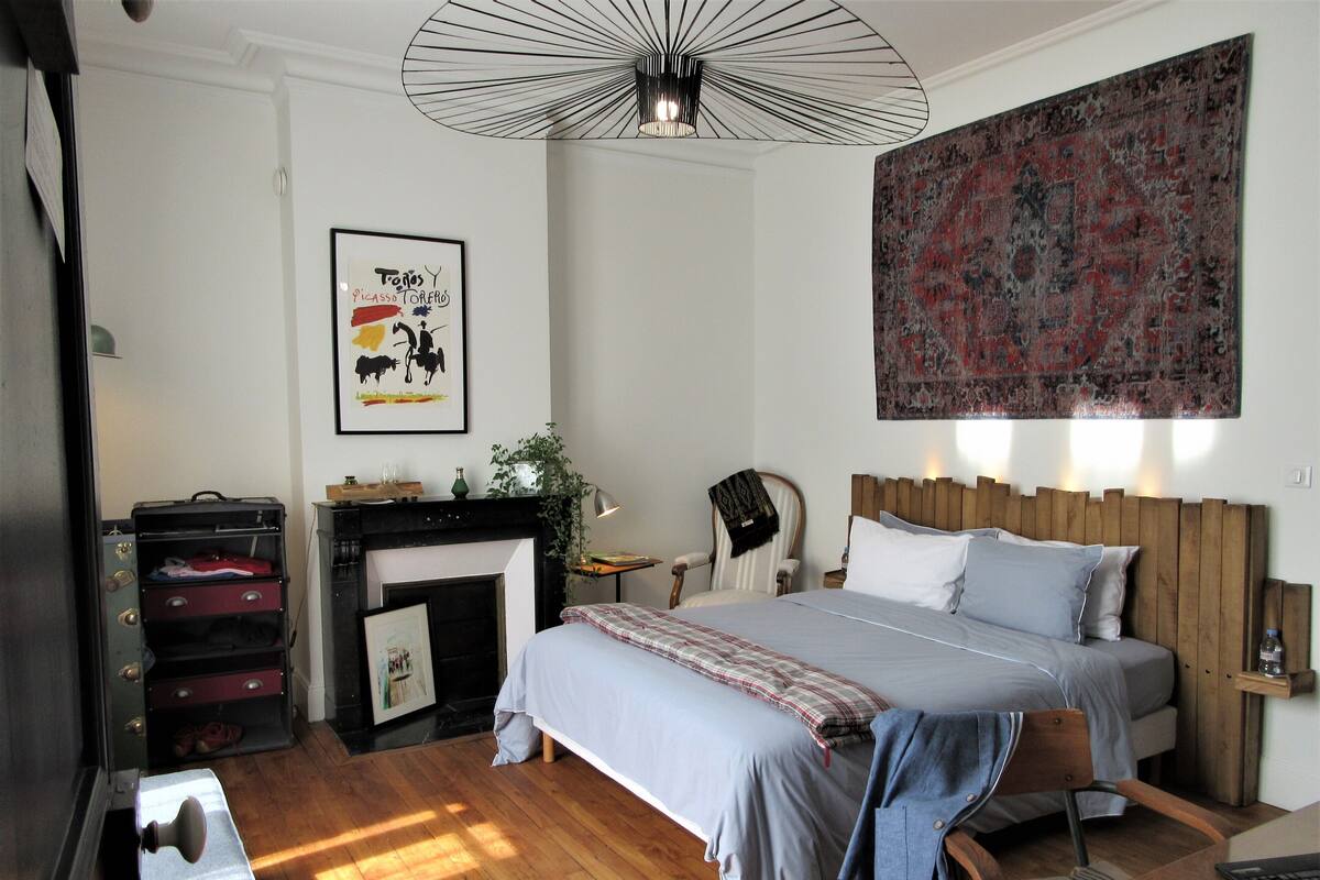 A bedroom with a bed, a chair, a desk, a fireplace, and a large tapestry.
