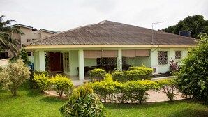A two-story house with a green roof, a porch with a brown awning, and a small garden with various plants. - Jayliz Lodge (Accra)