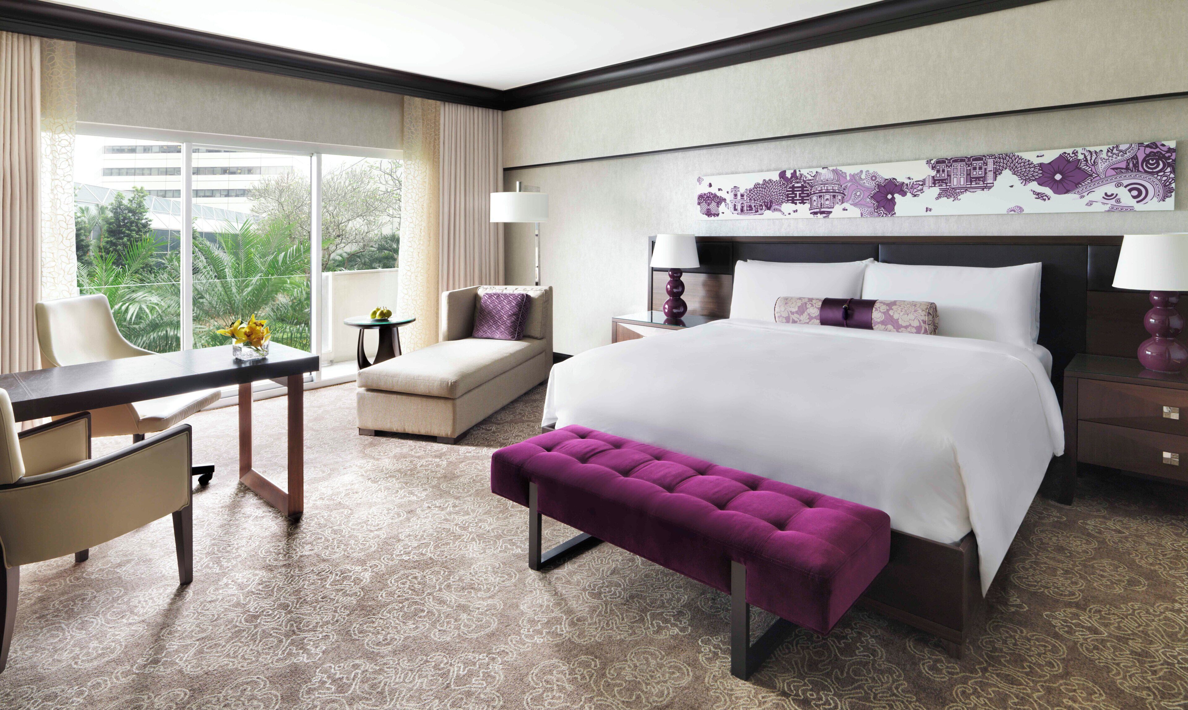 A hotel room with a large bed, a desk, a chair, a small table, and a view of greenery outside.