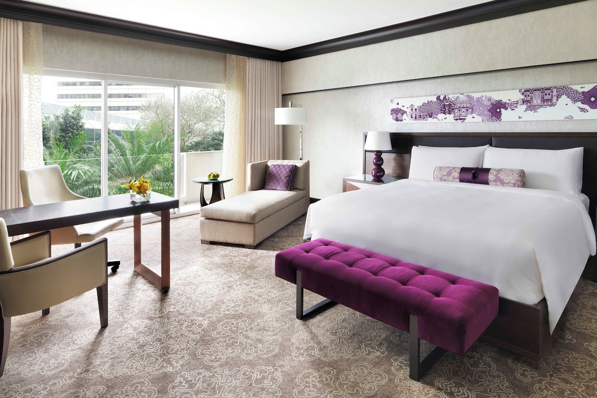 Une chambre d’hôtel avec un grand lit, un bureau, une chaise, une petite table et une vue sur la verdure par la fenêtre.