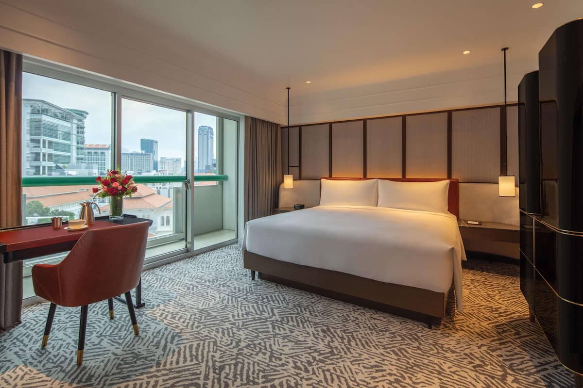 Een hotelkamer met een groot bed, een bureau met een stoel, een bloevas en uitzicht op de stad door grote ramen.