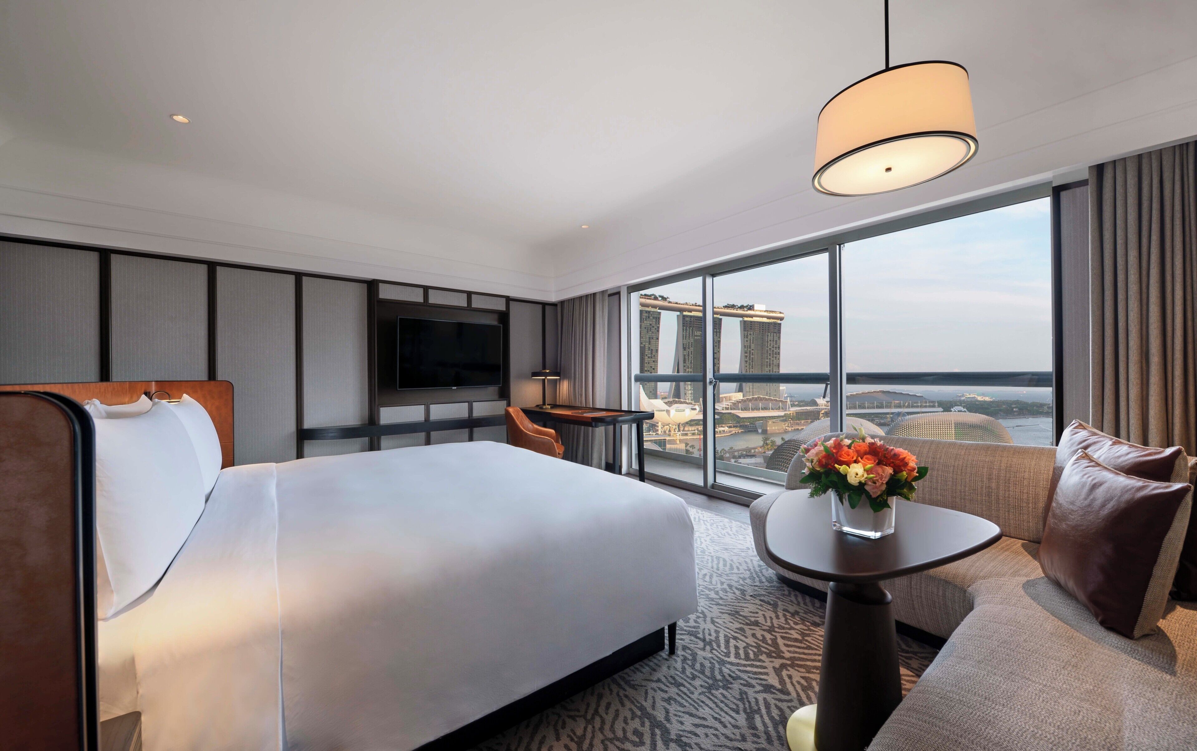 A hotel room with a bed, a TV, a desk, a chair, a small table with a vase, and a large window with a city view.