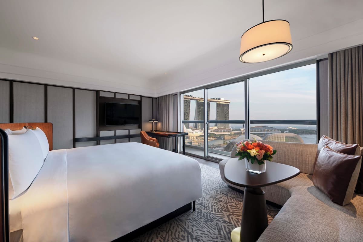 Een hotelkamer met een bed, een televisie, een bureau, een stoel, een klein tafeltje met een vaas, en een groot raam met uitzicht op de stad.