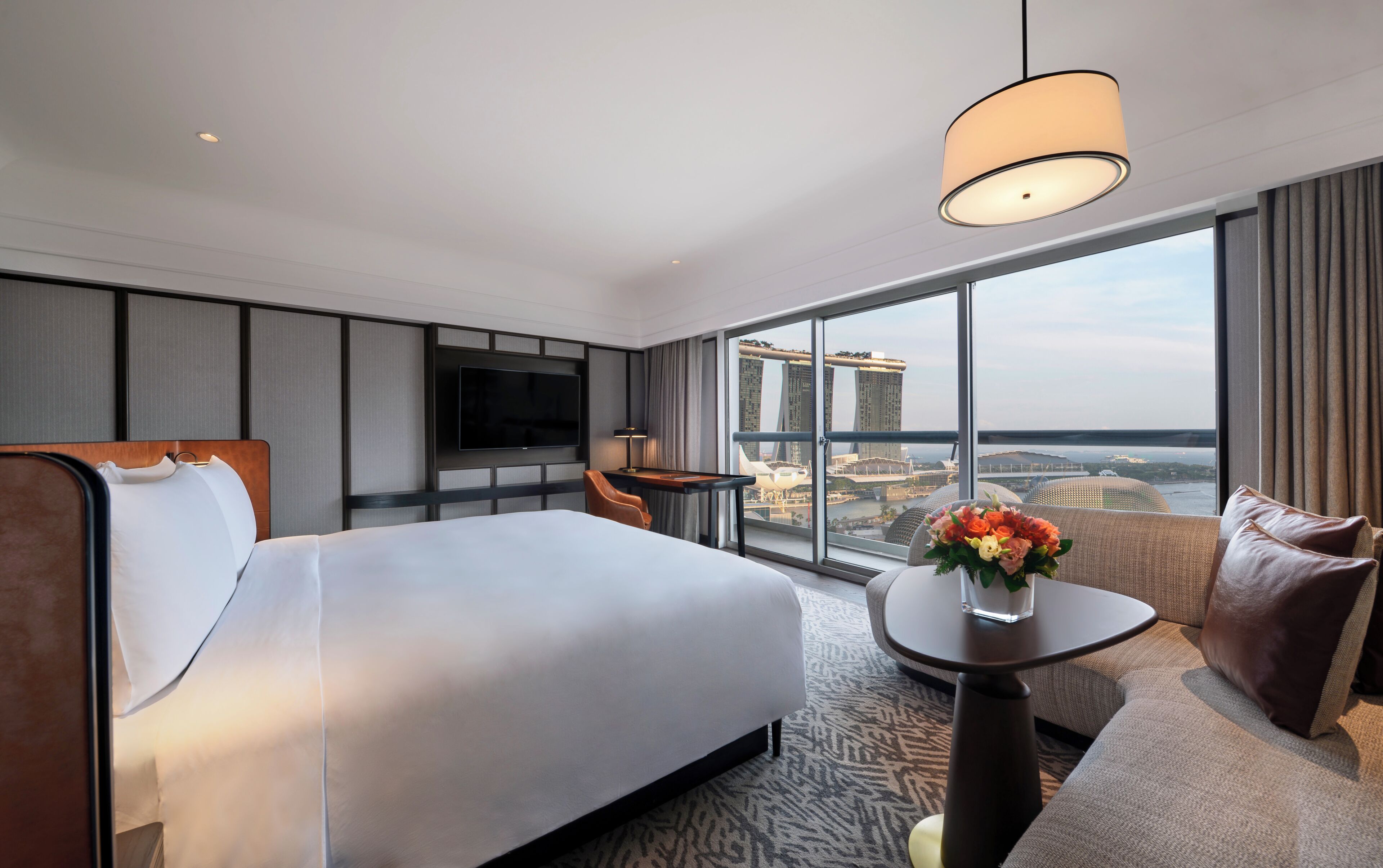 A hotel room with a large bed, a TV mounted on the wall, a small table, and a view of a cityscape through large windows.
