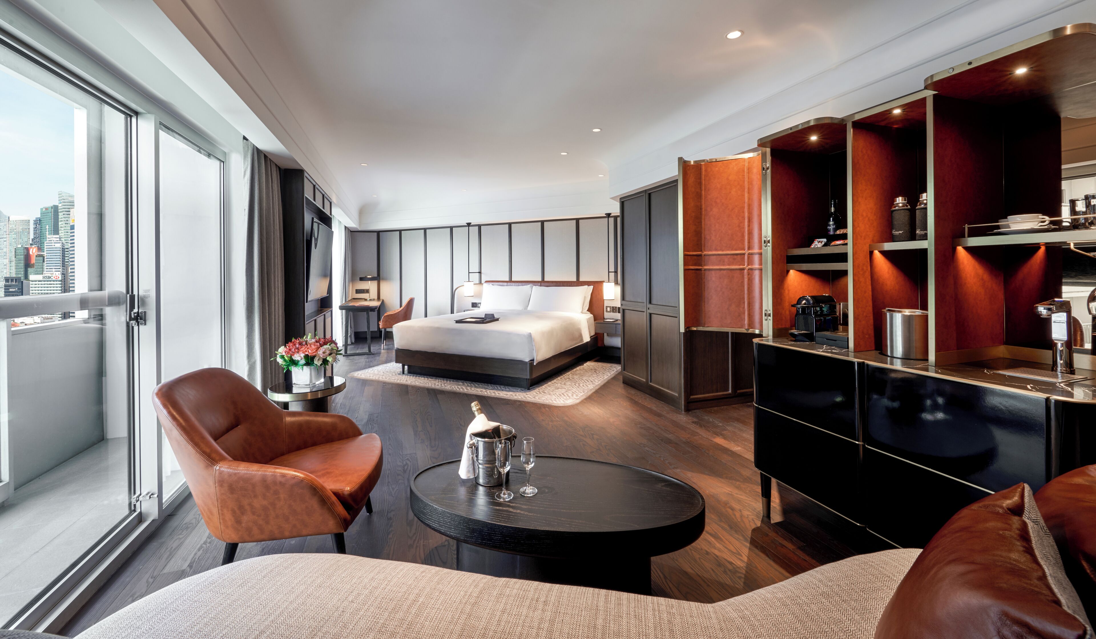 A modern hotel room with a large bed, a seating area with a coffee table, a minibar, and a city view.
