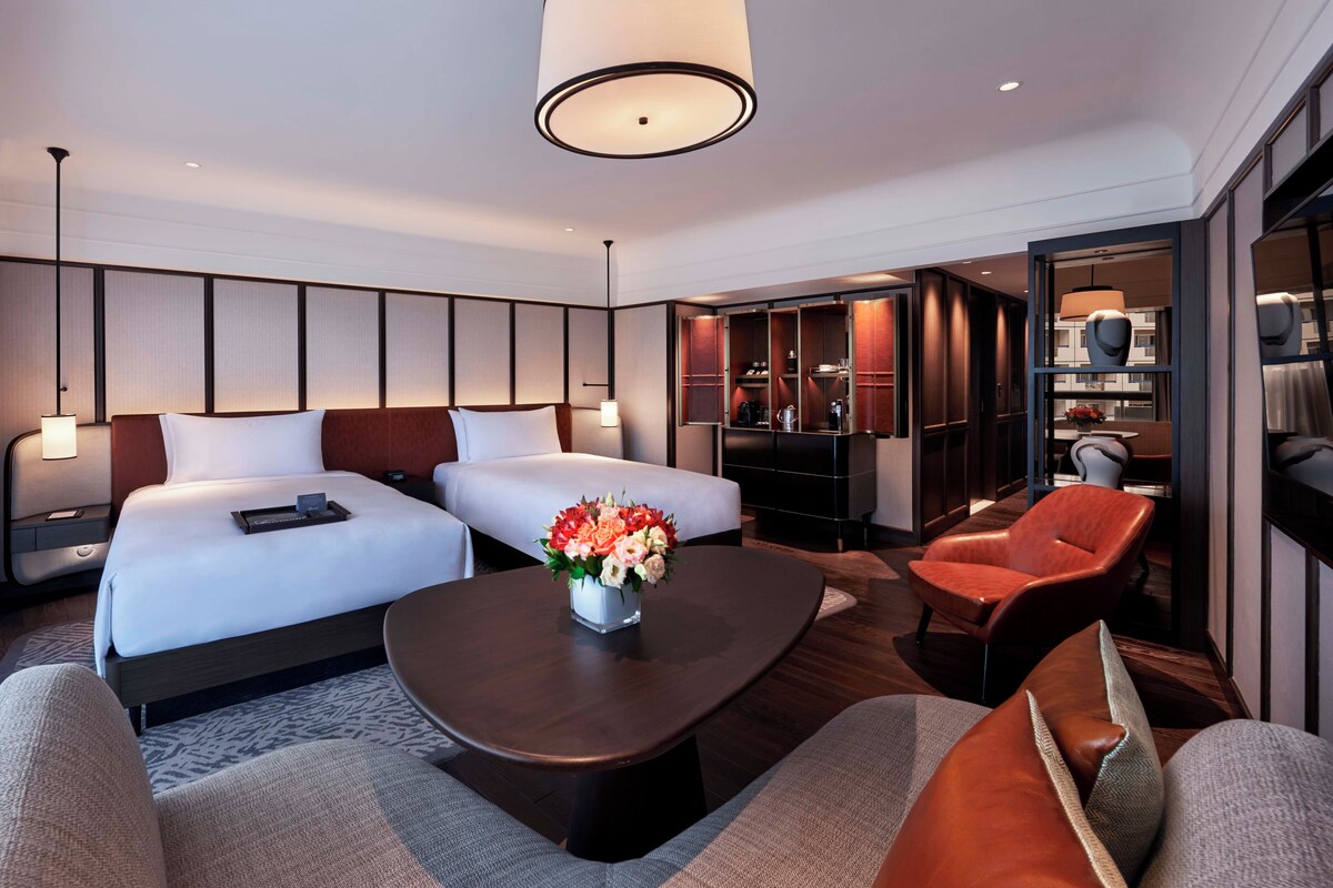 Une chambre d’hôtel avec deux lits, une table basse ornée d’un vase de fleurs, un canapé, un fauteuil rouge et un mini-bar.