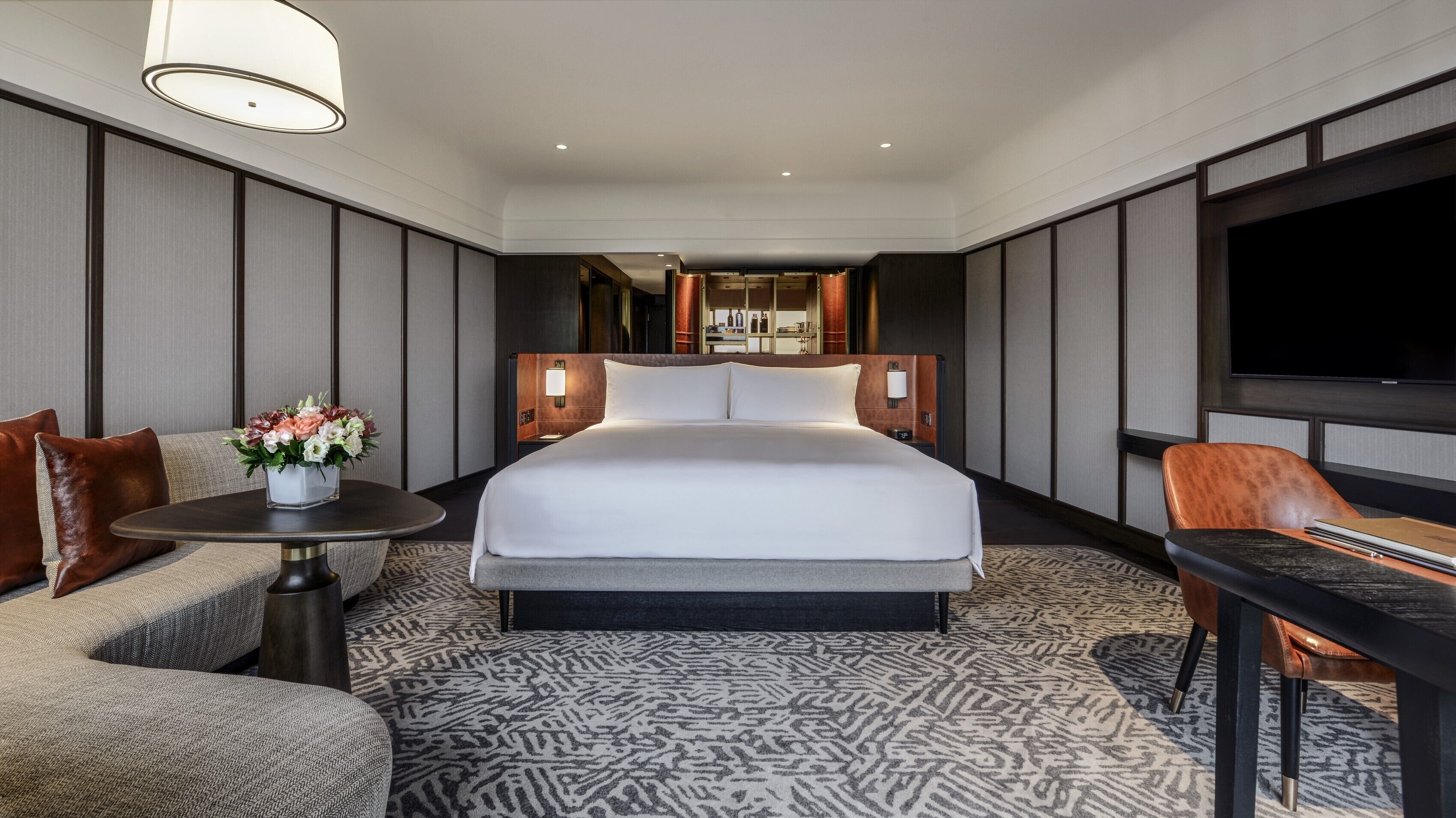 A hotel room with a large bed, a sofa, a small table with a vase of flowers, a TV, and a desk with a chair.