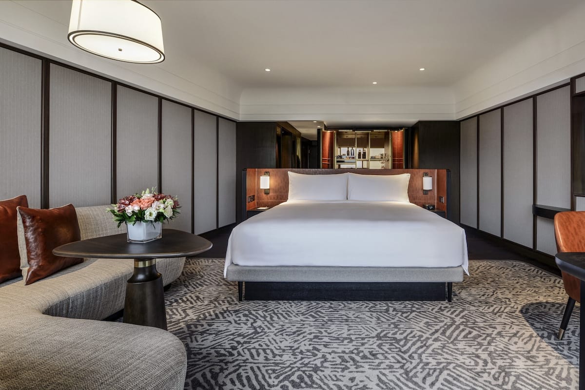 A hotel room with a large bed, a sofa, a small table with a vase of flowers, a TV, and a desk with a chair.