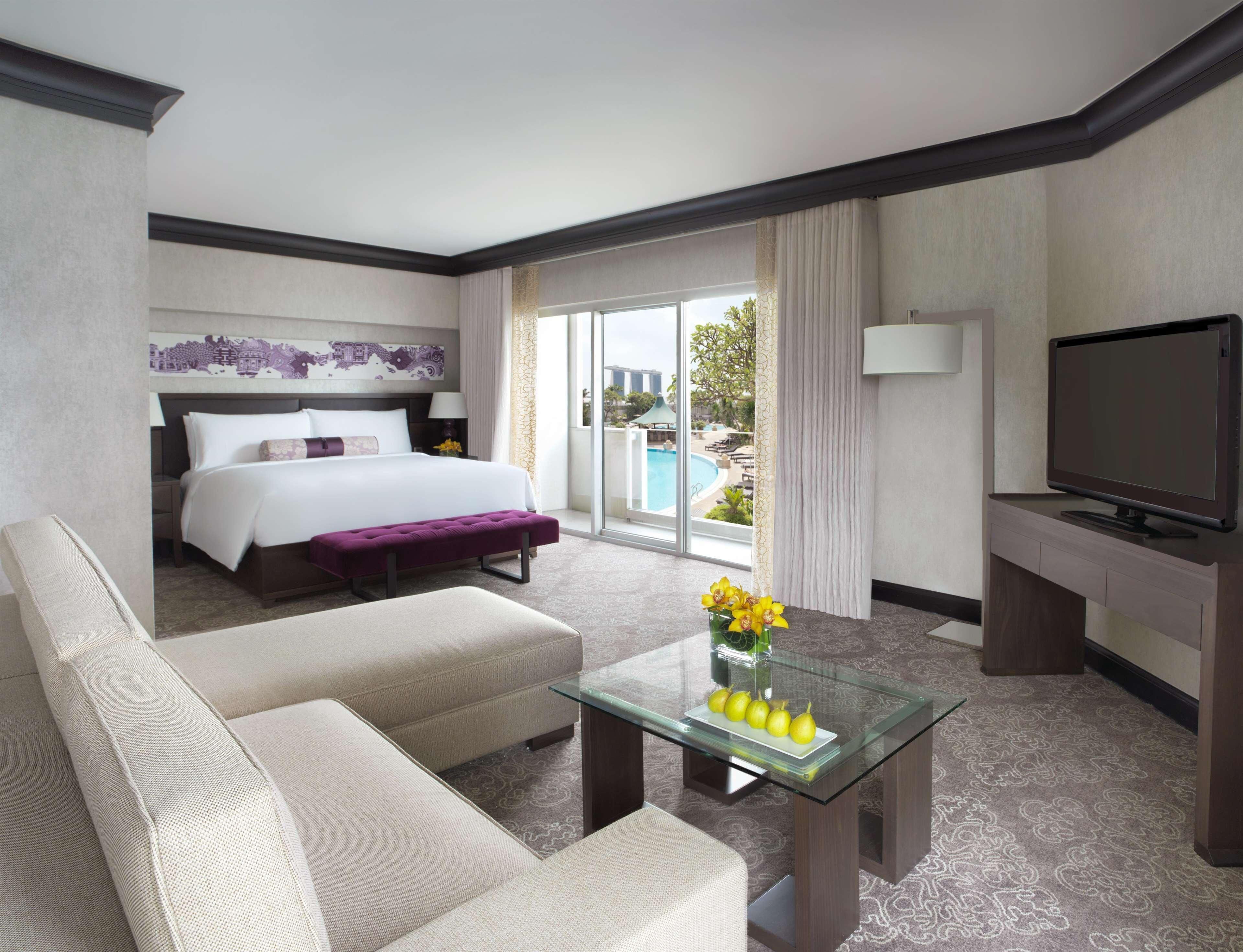 A hotel room with a bed, a sofa, a glass coffee table with fruit, a TV, and a view of a pool.