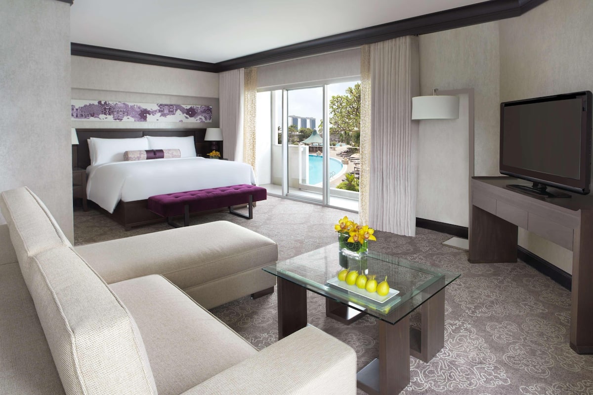 Hotelkamer met een bed, een bank, een glazen bijzettafeltje met fruit, een televisie en uitzicht op een zwembad.