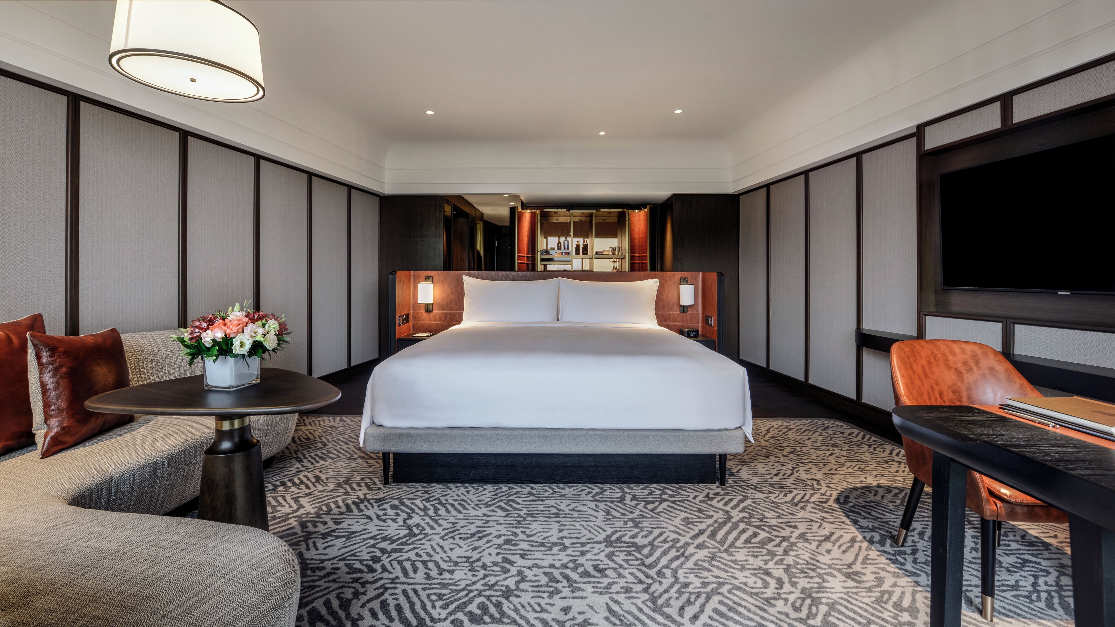 A hotel room with a large bed, a sofa, a small table with a vase of flowers, a TV, and a desk with a chair.