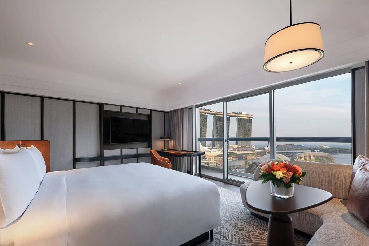 Hotelkamer met een groot bed, een televisie, een bureau, een stoel, een klein tafeltje met een vaas en uitzicht op een stadsbeeld door grote ramen.