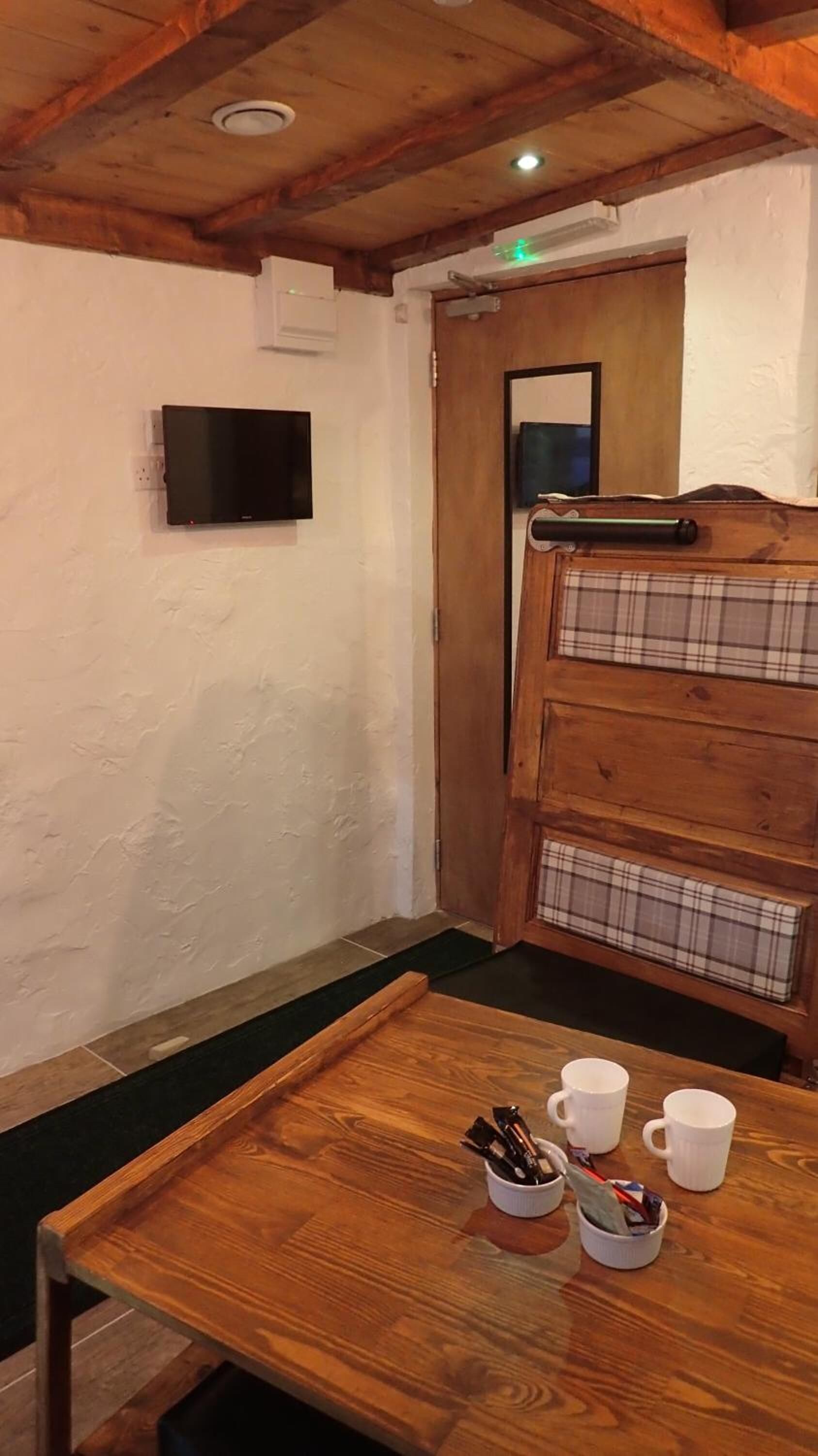 A wooden table with two white mugs and a cup of coffee, a plaid bench, and a television mounted on the wall.