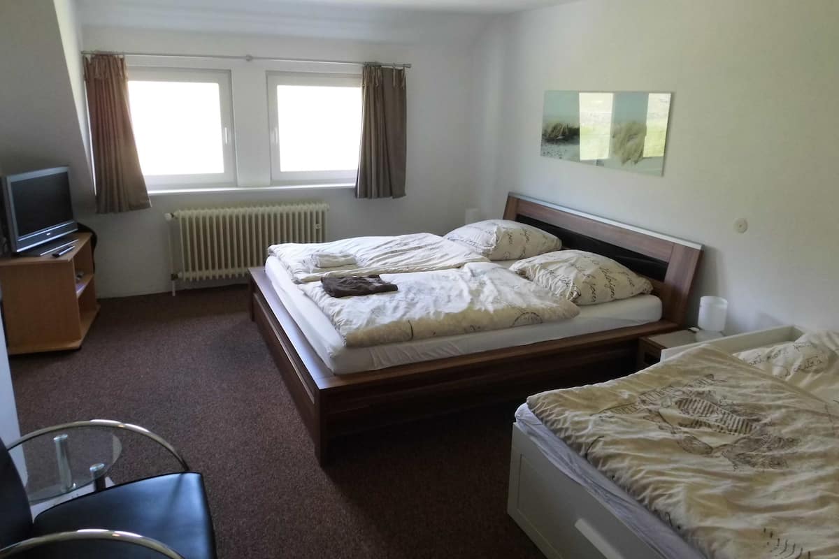 Une chambre d’hôtel avec un lit simple, un deuxième lit, un bureau avec une chaise, une télévision et une fenêtre avec des rideaux.