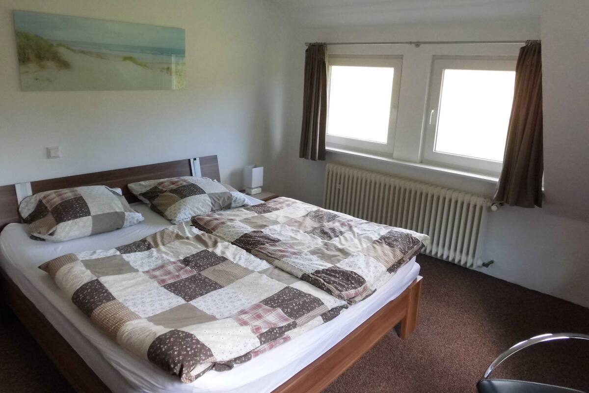 Un lit double avec une couvre-lit à carreaux, une table de chevet avec une lampe, une fenêtre avec des rideaux et un tableau accroché au mur.