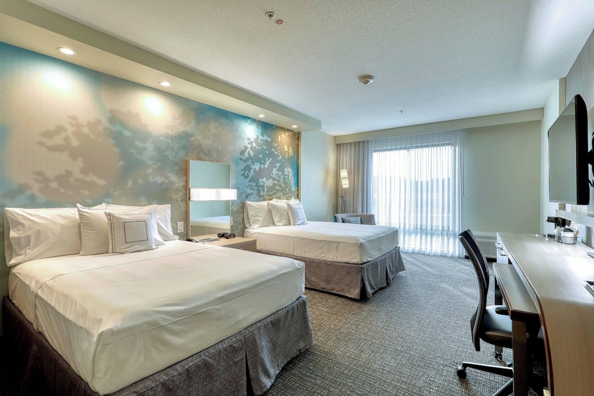 A hotel room with two beds, a desk, a chair, a large mirror, and a window with curtains.