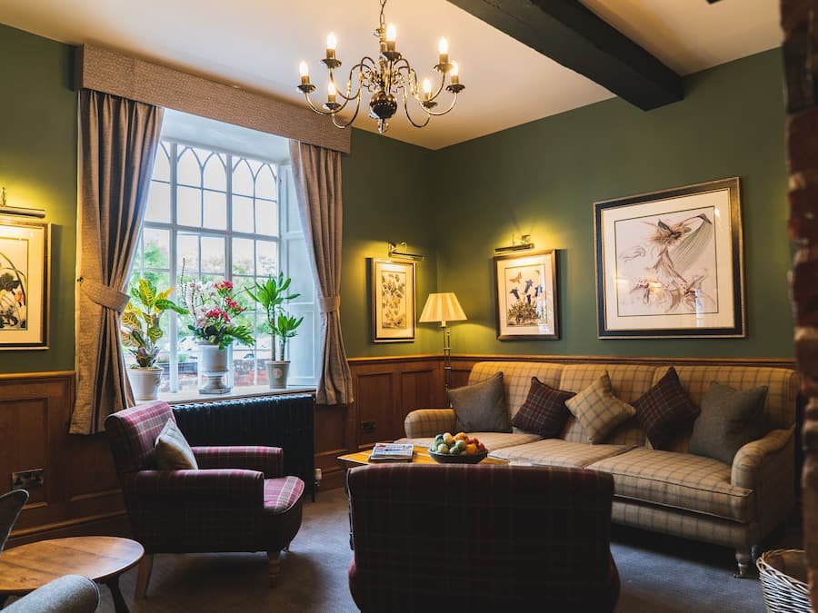 A cozy living room with a checkered sofa, a plaid armchair, and a table with a fruit bowl. There are framed pictures on the walls, a window with curtains, and a chandelier.