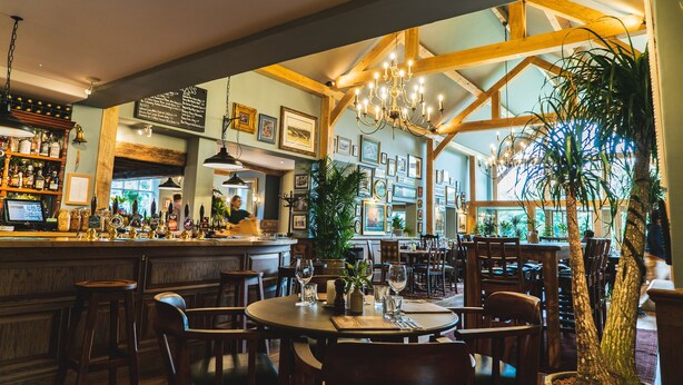 Un restaurant avec des poutres en bois, un coin bar avec des bouteilles exposées et des tableaux encadrés aux murs.