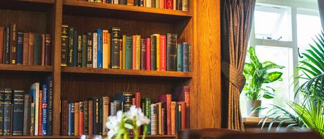 Una biblioteca con estanterías de madera llenas de libros, una mesa con un jarrón de flores y una planta en maceta cerca de la ventana.