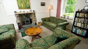A cozy living room with patterned green sofas, a fireplace, a bookshelf, and a window with a view of greenery. - Cuffern Manor (Haverfordwest)