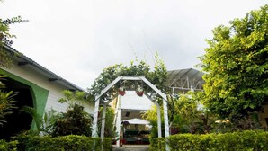 A pathway leading to a covered area with a white archway, surrounded by lush greenery and a building with a green roof.