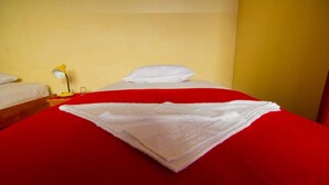A hotel room with a bed covered in a red blanket and white sheets, a yellow lamp on a bedside table, and a plain yellow wall.