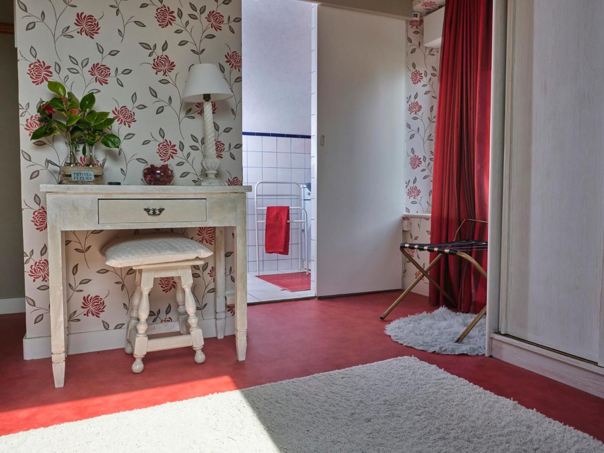 Une pièce avec un bureau de style vintage, un petit tabouret, une plante en pot, une lampe et un rideau rouge.