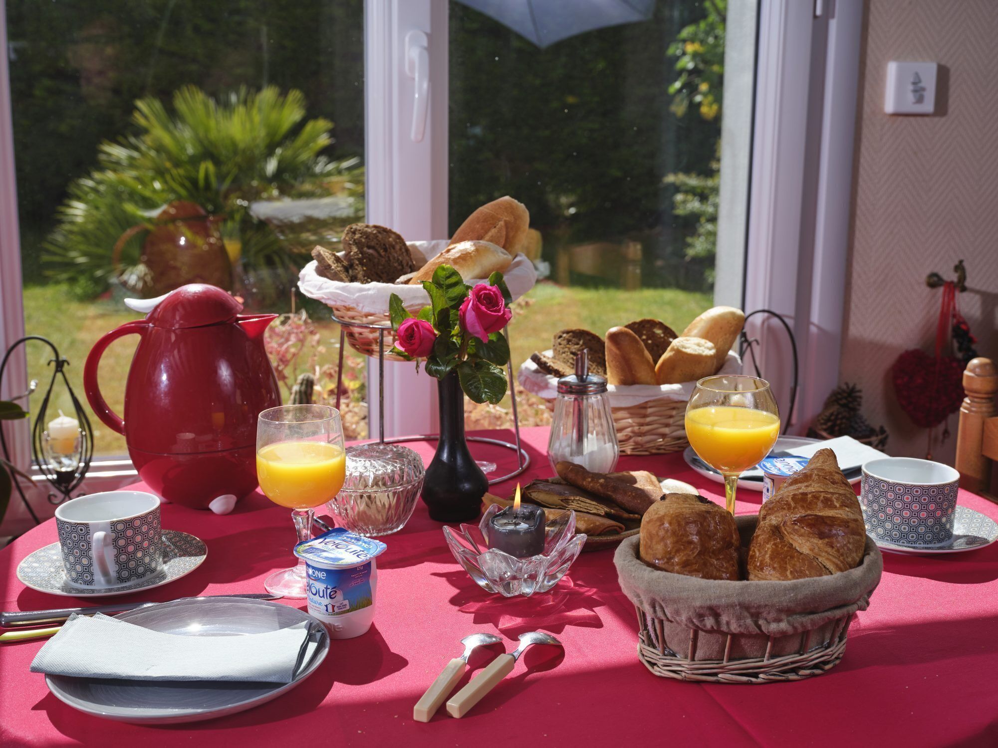 Une table de petit-déjeuner avec une nappe rouge, une bouilloire rouge, un vase rempli de fleurs roses, un panier de pain et un verre de jus d’orange.