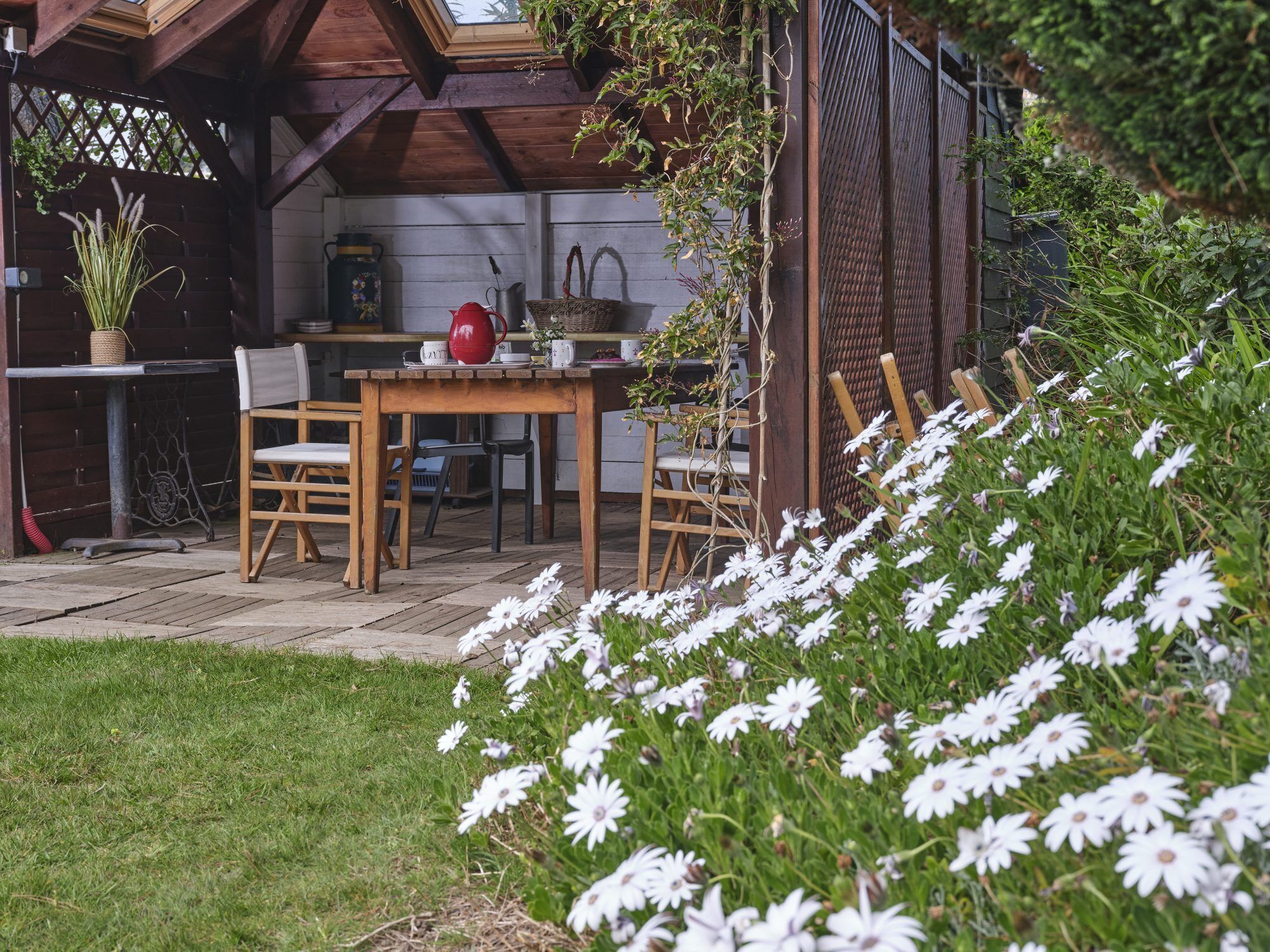 A wooden outdoor dining area with a table and chairs, surrounded by a garden with white flowers.