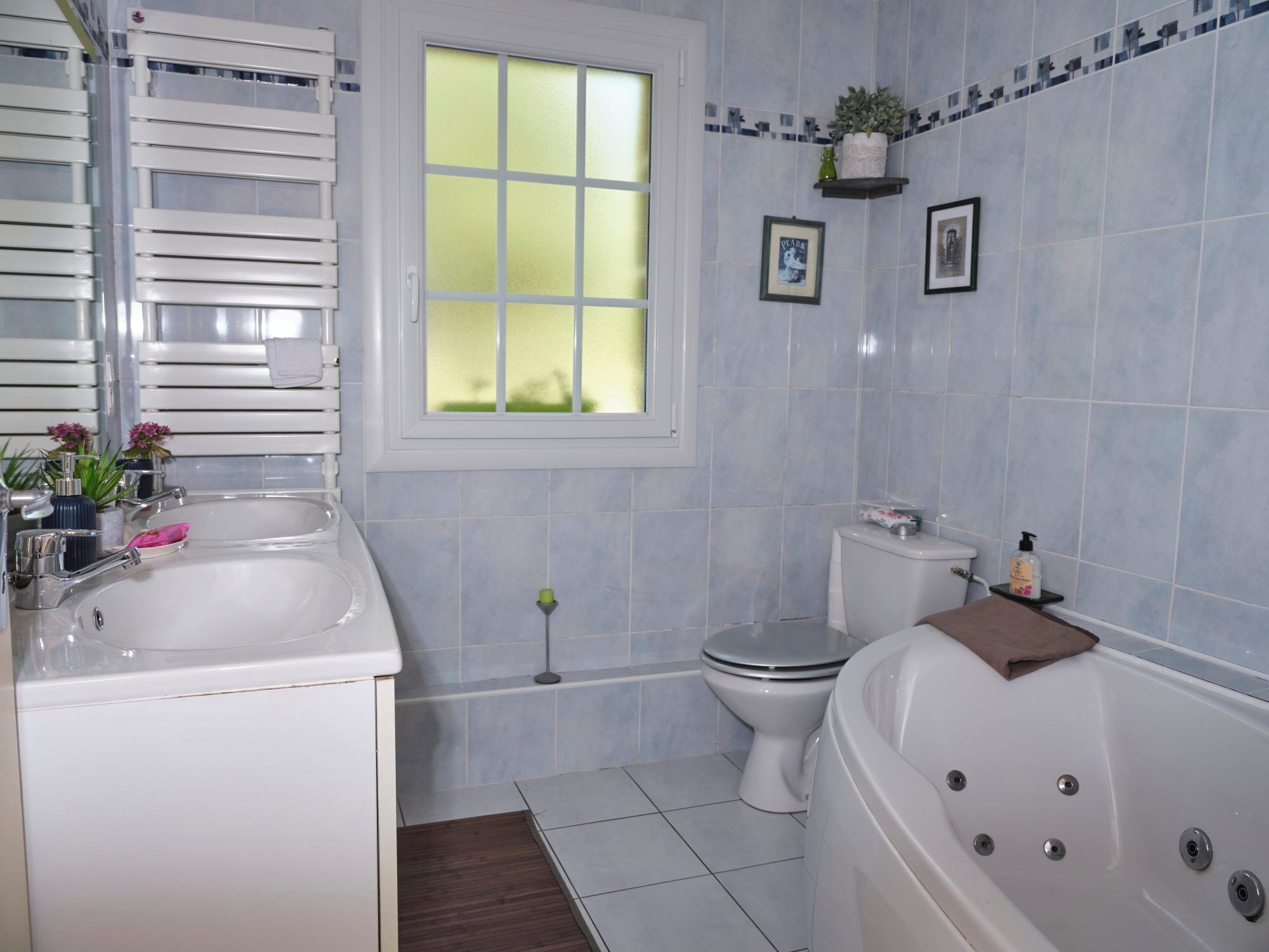 Un bagno con un lavandino bianco, un water e una vasca da bagno. Si vede una finestra con vetro smerigliato, una pianta in vaso sulla parete e un asciugamano nella vasca.
