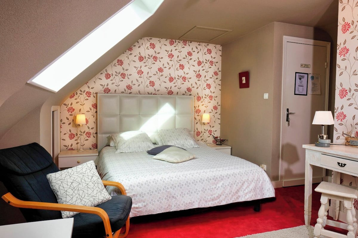 A bedroom with a sloped ceiling, floral wallpaper, a bed with white bedding, a nightstand with a lamp, a chair, and a desk with a lamp.