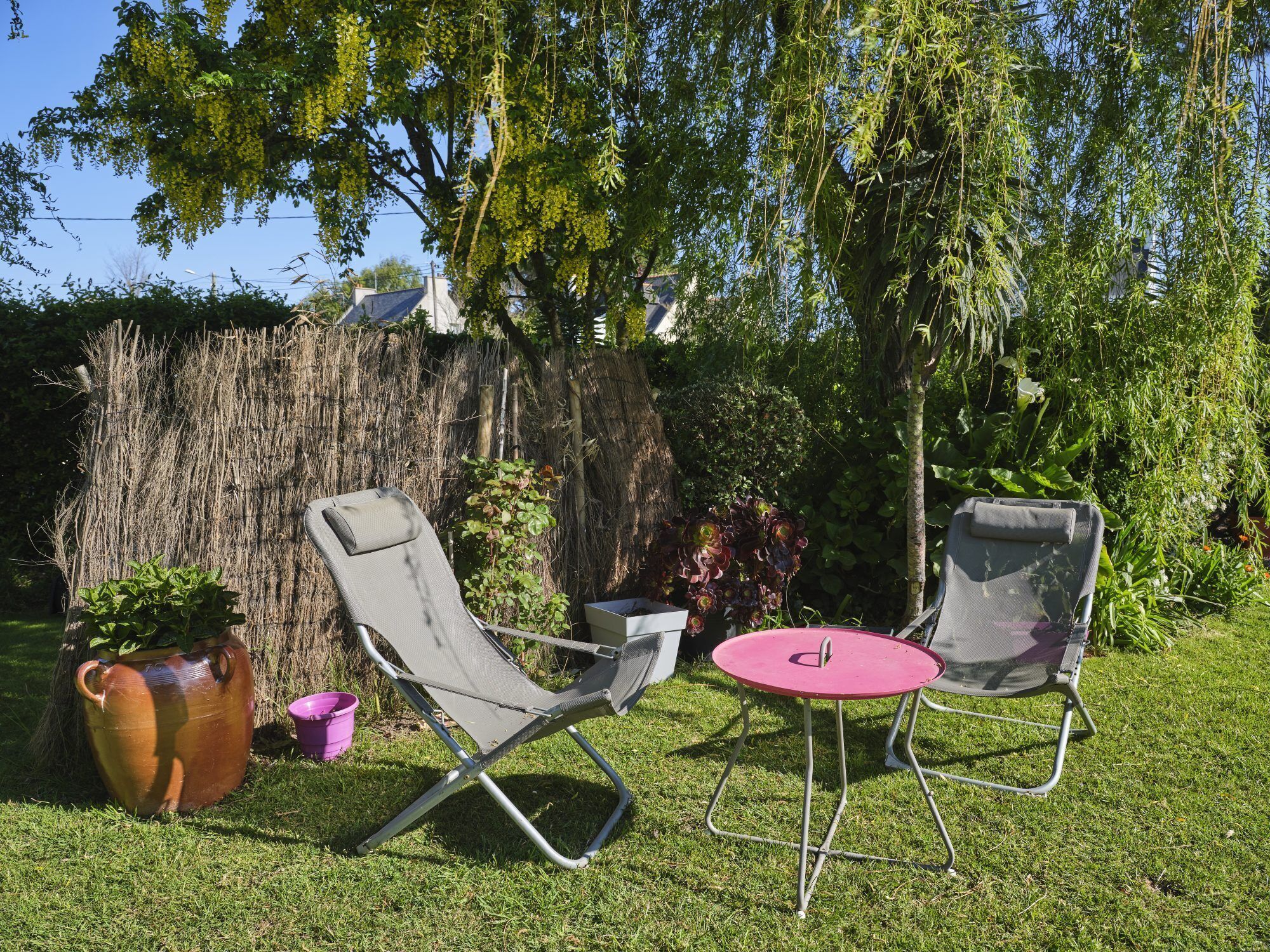 Un espace extérieur aménagé avec deux chaises et une petite table, entouré de verdure et bordé d’un mur recouvert de chaume.
