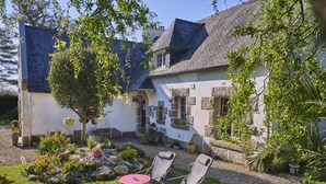 A stone house with a sloped roof, a small garden with a lawn, and two chairs. - Les Roses (Santec)