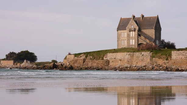 A stone building with a steep roof, located near a rocky shoreline. - Les Roses (Santec)