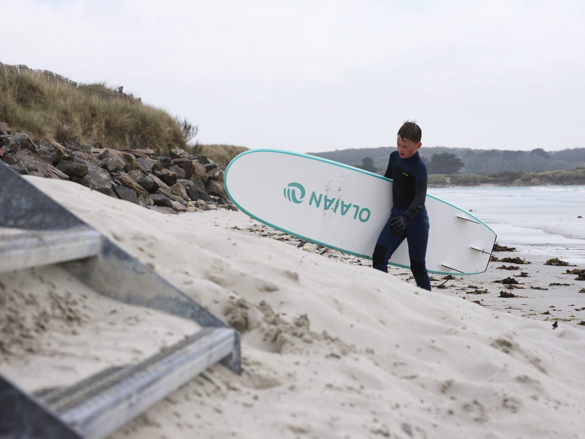 Una persona che trasporta un'asse da surf con il marchio "O'LIAN" su una spiaggia sabbiosa.