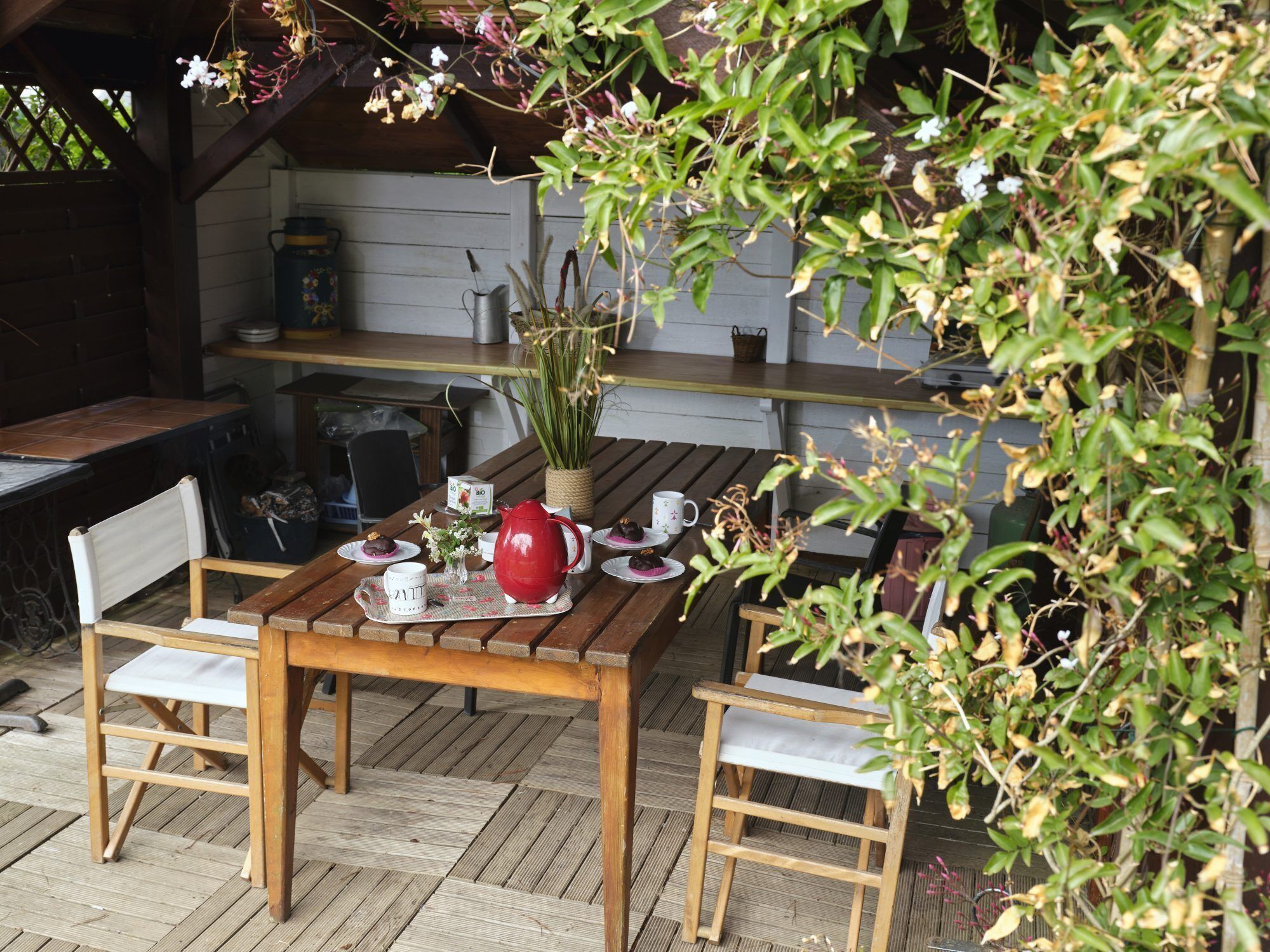 Une table en bois placée à l’extérieur, avec des chaises, une bouilloire rouge et un petit vase blanc contenant des fleurs.