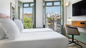 A hotel room with two beds, a desk, a chair, a TV, and large windows with a city view. - One Shot Aliados Goldsmith  (Porto)