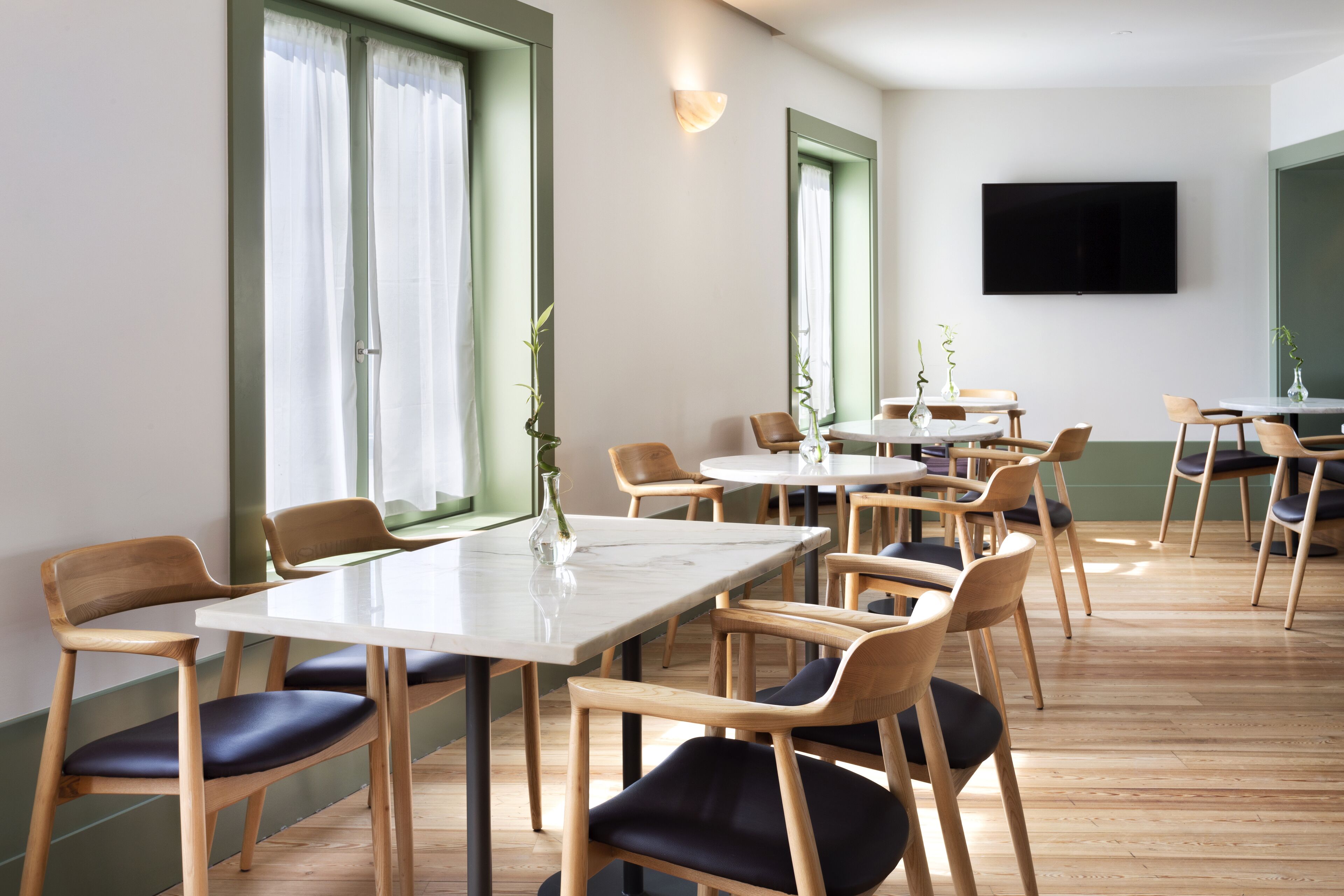 Un comedor moderno con sillas y mesas de madera, ventanas grandes con cortinas y un televisor de pantalla plana en la pared.