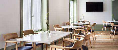 Un comedor moderno con sillas y mesas de madera, ventanas grandes con cortinas y un televisor de pantalla plana en la pared.