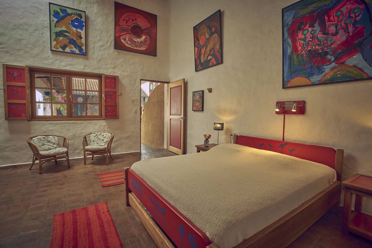Een slaapkamer met een groot bed, twee stoelen, een raam met luiken en verschillende schilderijen aan de muur.