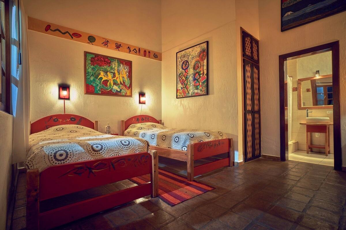 Een kamer met twee bedden, elk voorzien van een bed met een patroon, een rood hoofdeinde en een klein nachtkastje. Twee wandlampen, een houten deur met decoratief paneel, en een zichtbare badkamer door een open deur.