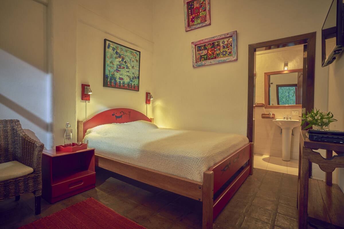 Een slaapkamer met een houten bed, een rode nachtkastje, een rieten fauteuil, een rood tapijt, een aan de muur bevestigde televisie en een badkamer met een wastafel en spiegel.
