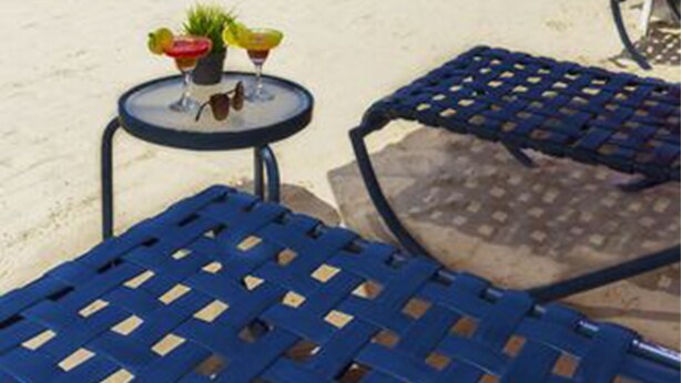 Eine Strandkulisse mit einem Tisch unter einem roten Sonnenschirm, einem blauen Stuhl und Blick auf das Meer.