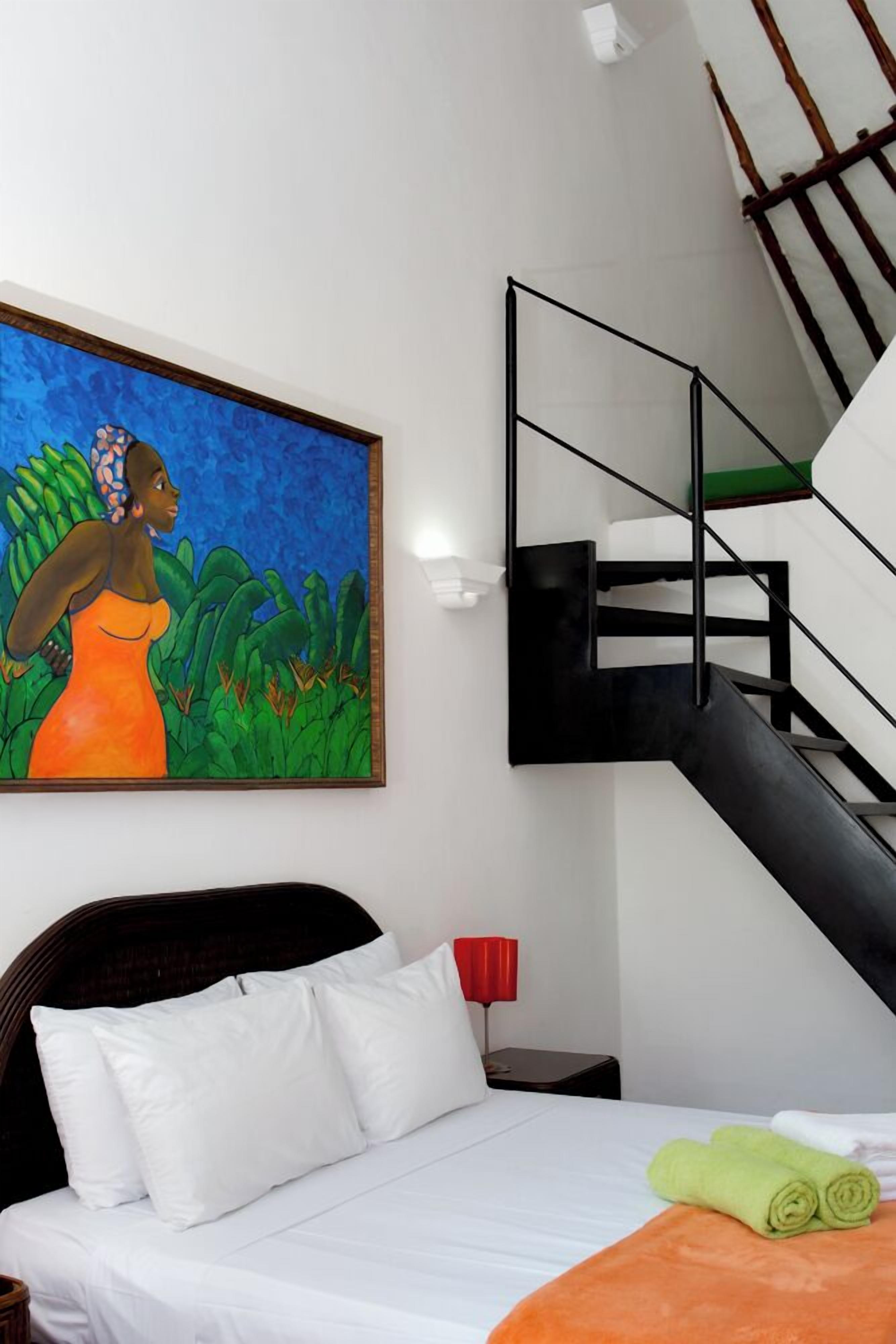 A hotel room with a bed, a painting, a staircase, and a nightstand with a red lamp.
