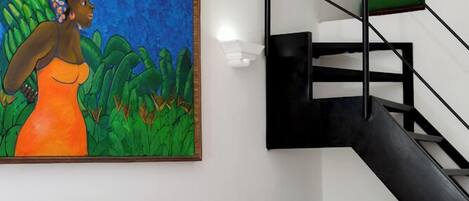 A hotel room with a bed, a painting, a staircase, and a nightstand with a red lamp.